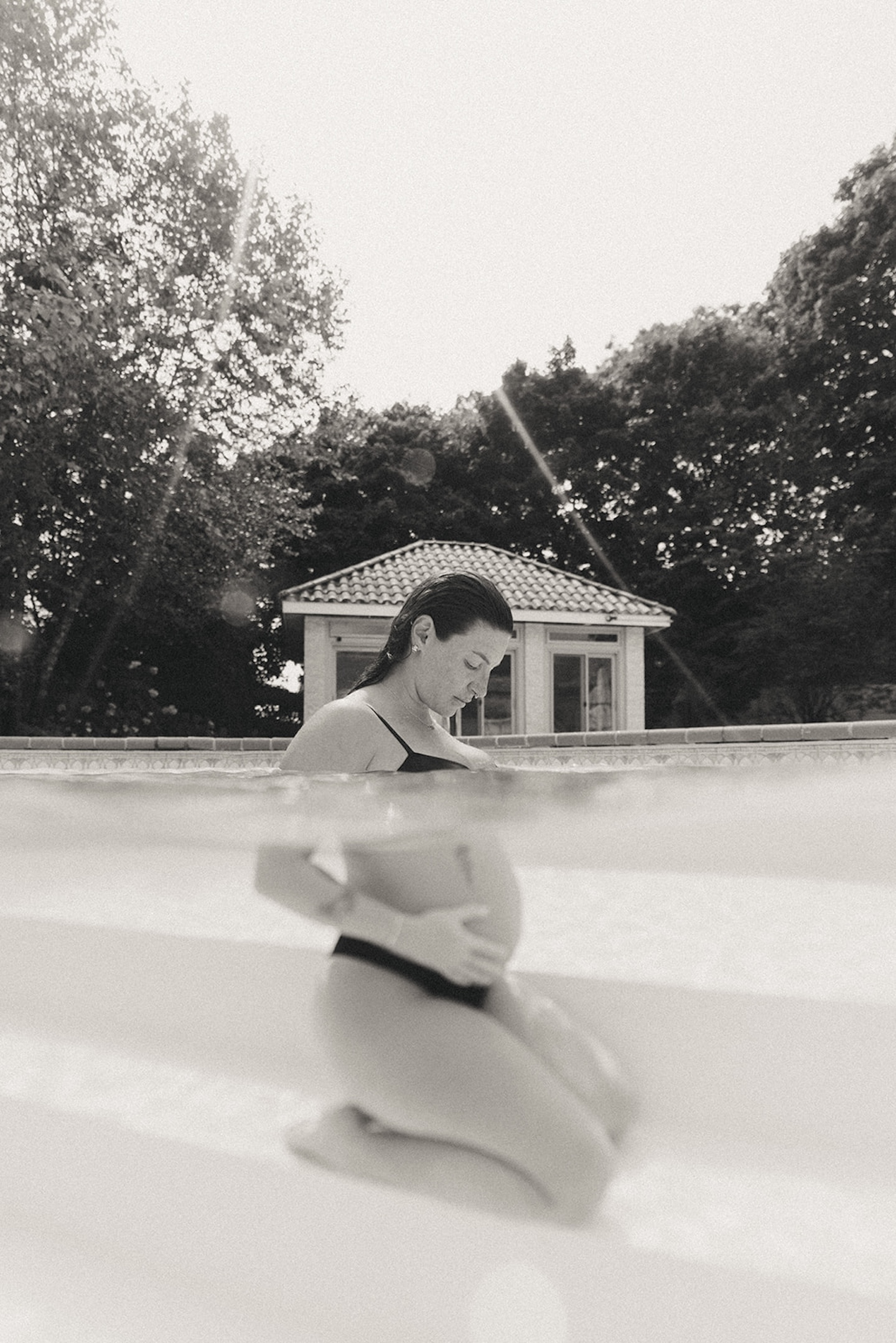 Black and white water maternity picture shot at waterline, pregnant woman standing in pool gazing downward, pool house and trees softly blurred in background — emotional above-water pregnancy photo by maternity photographer, Elsie Goodman.