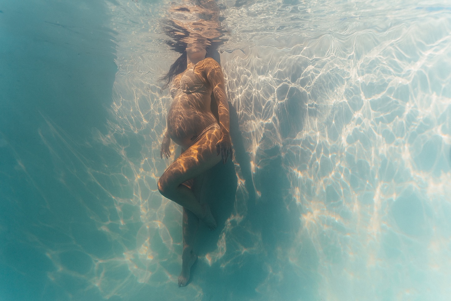 Full body underwater maternity photography of a pregnant woman in white bikini, suspended in clear turquoise pool water with shimmering light rays by maternity photographer, Elsie Goodman.