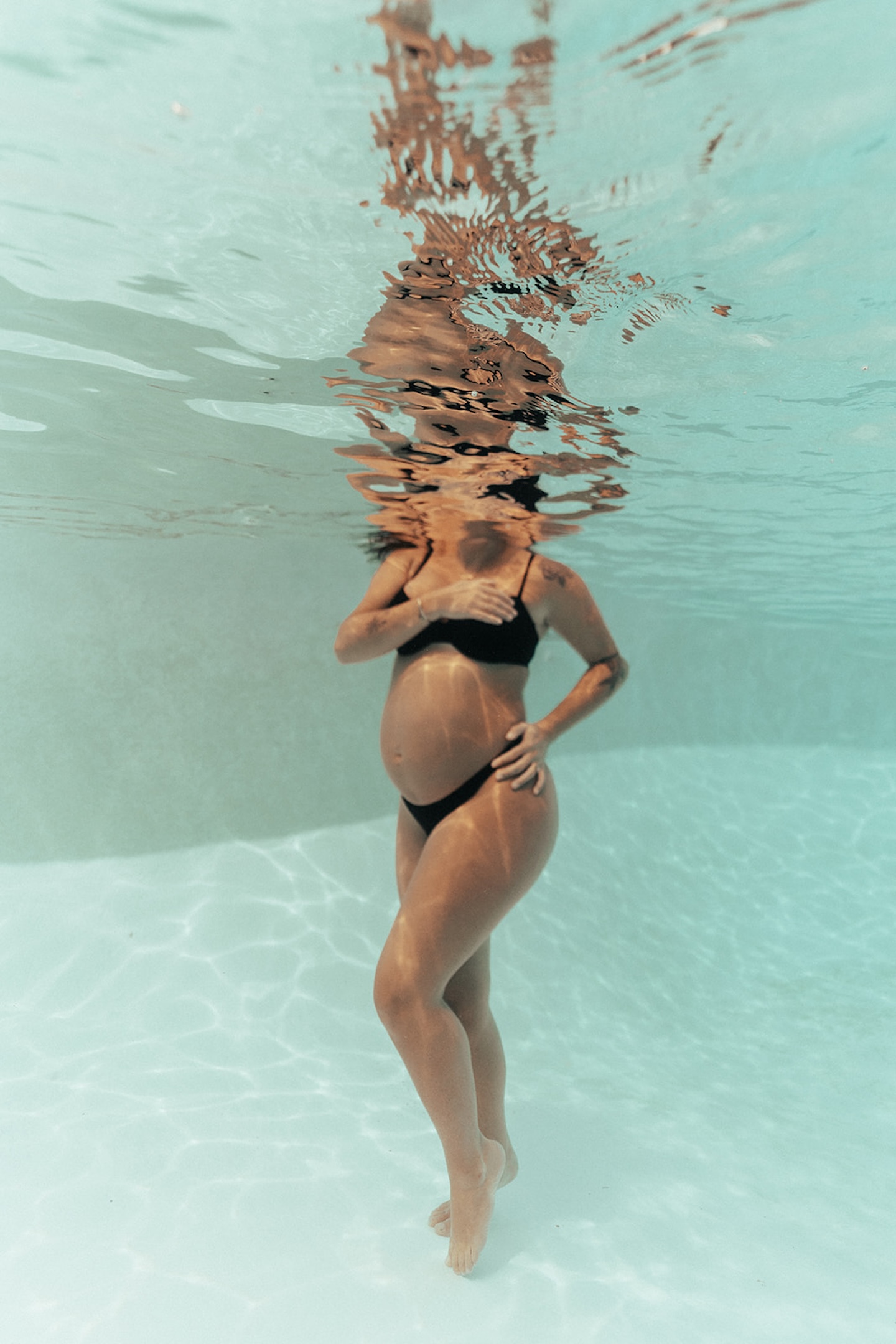 Underwater pregnancy photo of expectant mother in black bikini standing on pool floor, hands cradling belly, turquoise water surrounding her — water maternity picture with warm reflections at surface by maternity photographer, Elsie Goodman.