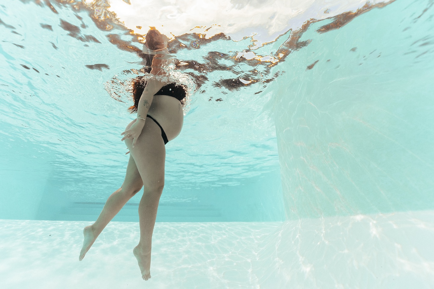 Underwater maternity picture of pregnant woman with long dark hair arching back gracefully, cradling bump, sunlight dappling through turquoise pool water — serene underwater pregnancy photo by maternity photographer, Elsie Goodman.