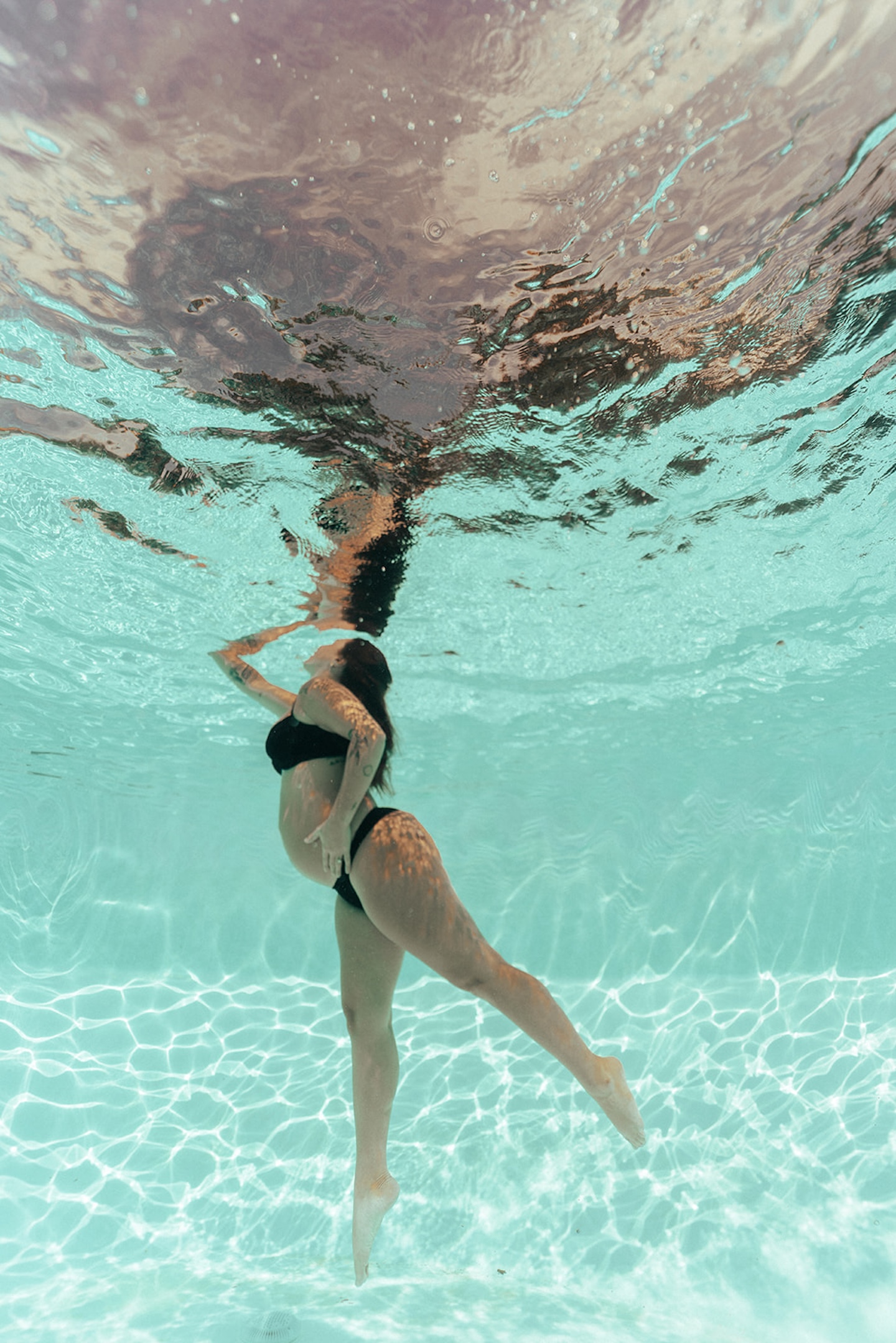 Underwater pregnancy photo of side-profile pregnant woman in black bikini, hair floating, reaching upward through purple-tinted turquoise pool water — moody water maternity picture by maternity photographer, Elsie Goodman.