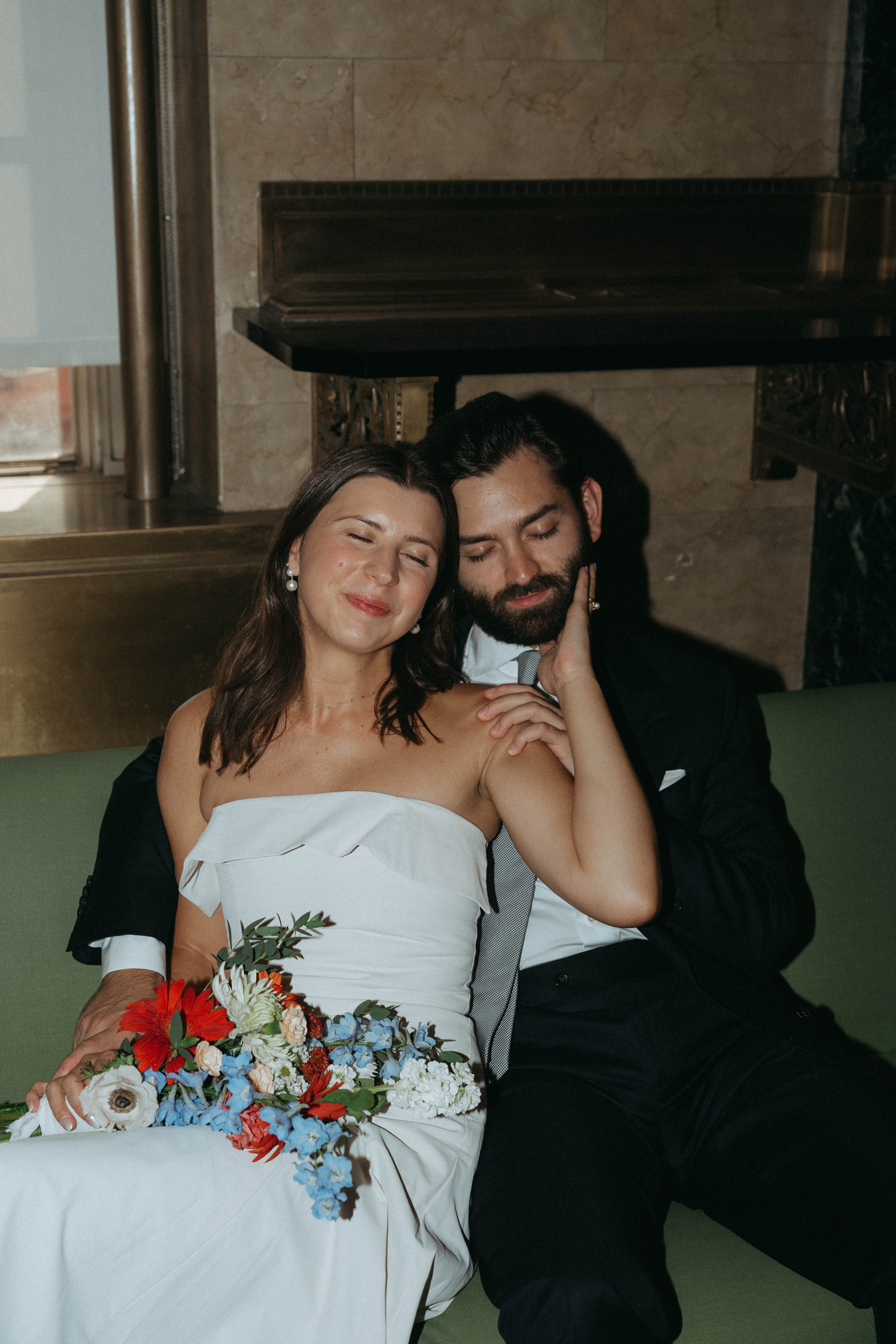 Bride and groom with a red bouquet on a bench inside the NYC City Hall marriage bureau