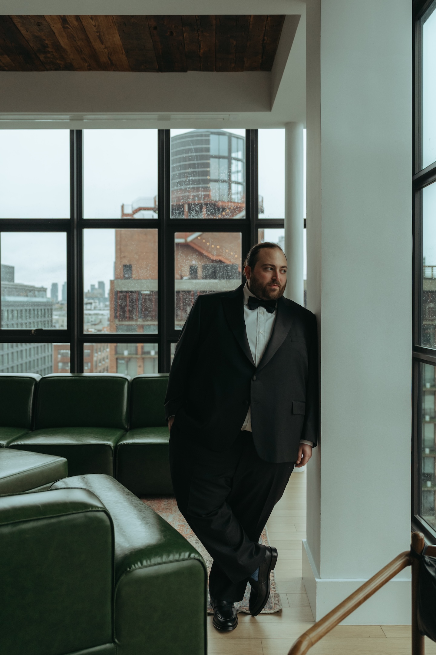 Groom in a tuxedo by a green sofa and city-view windows before his NYC elopement