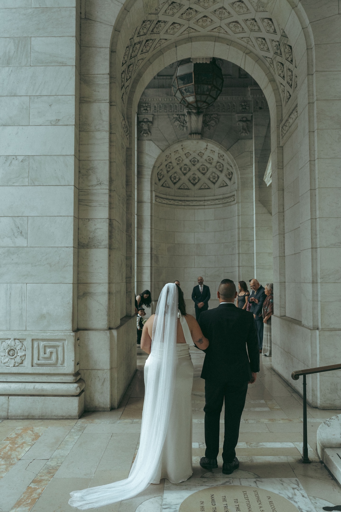 Bride in a long veil and groom in a suit walking under the New York Public Library arch for their elopement ceremony