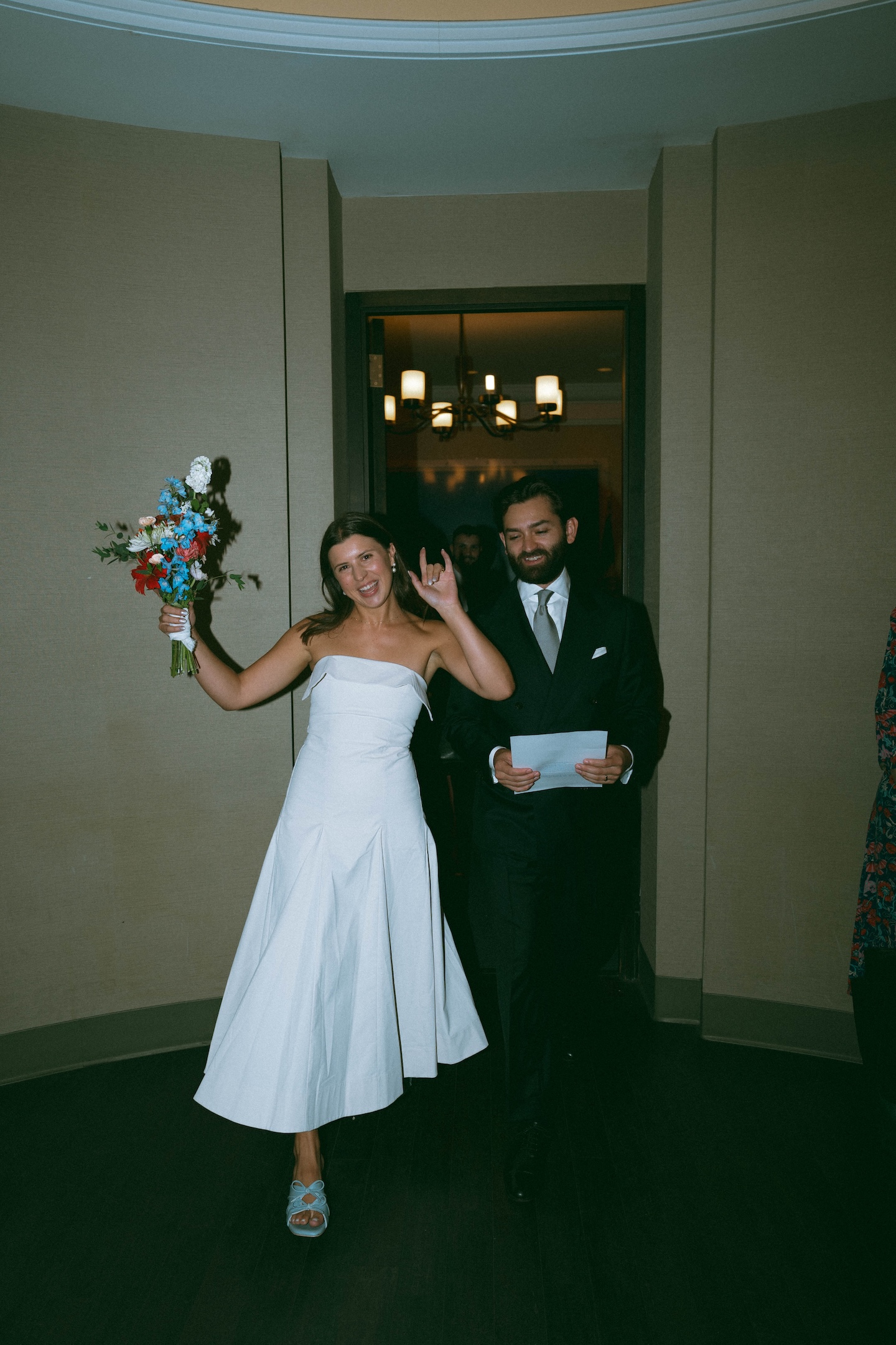 Bride holding a red and blue bouquet walking the City Hall corridor with her groom holding marriage certificate after eloping