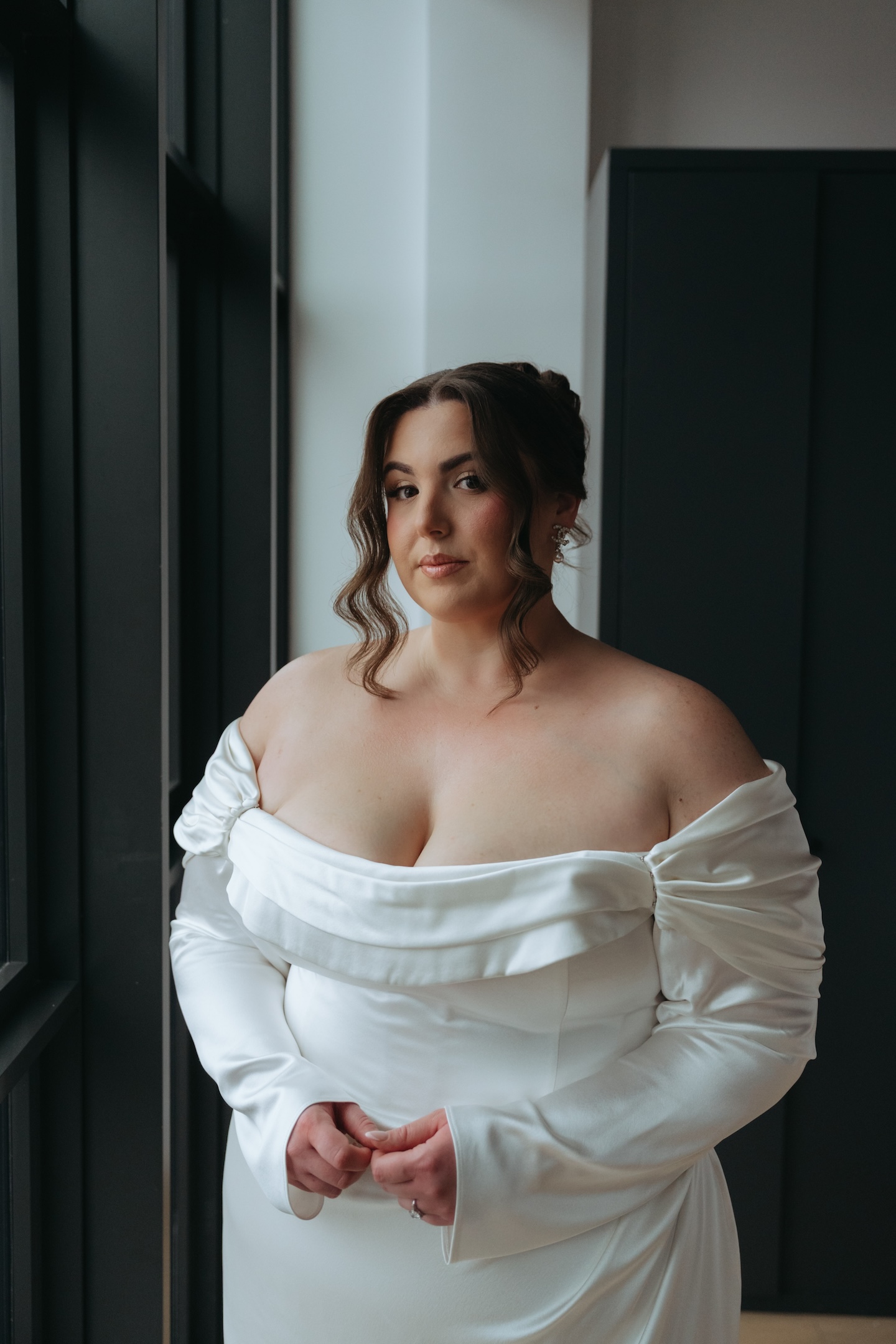 Bride in an off-shoulder white gown by floor-to-ceiling windows on her elopement day